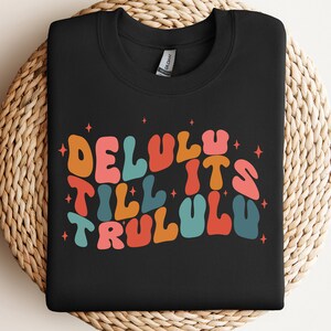 Puede incluir: Sudadera negra con un gr&aacute;fico colorido que dice "Delulu Till It's Trululu" en una fuente retro.