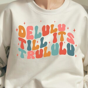 Puede incluir: Sudadera blanca con un estampado gr&aacute;fico colorido que dice "Delulu till it's trululu" con estrellas alrededor del texto.