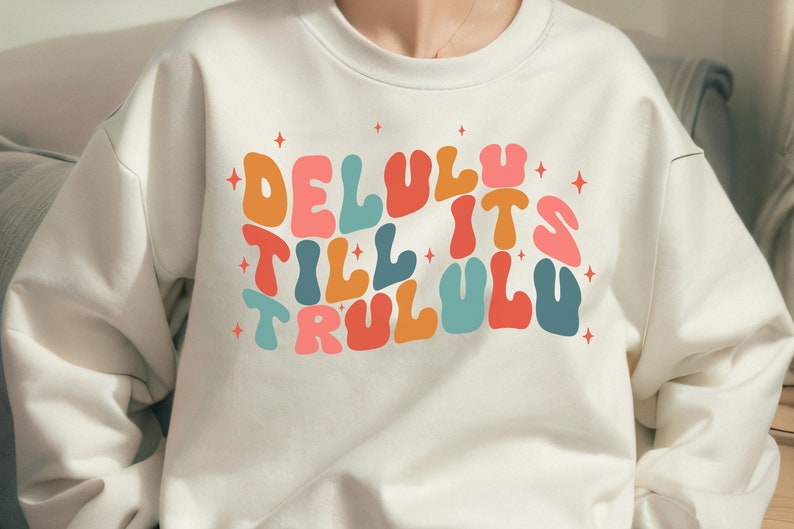 Puede incluir: Sudadera blanca con un estampado gr&aacute;fico colorido que dice "Delulu till it's trululu" con estrellas alrededor del texto.