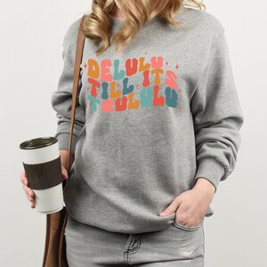 Puede incluir: Una sudadera gris con el texto "Delulu till it's trulu lu" en letras de colores de estilo retro.