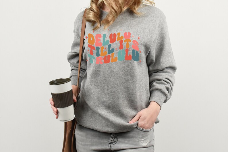 Puede incluir: Una sudadera gris con el texto "Delulu till it's trulu lu" en letras de colores de estilo retro.