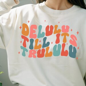 Puede incluir: Una sudadera blanca con un dise&ntilde;o gr&aacute;fico colorido que dice "Delulu Till It's Trululu" con estrellas alrededor del texto.