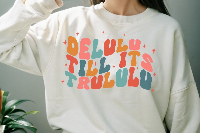 Puede incluir: Una sudadera blanca con un dise&ntilde;o gr&aacute;fico colorido que dice "Delulu Till It's Trululu" con estrellas alrededor del texto.