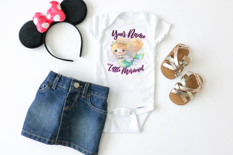 May include: A white bodysuit with a mermaid illustration and the text "Your Name the Little Mermaid". The bodysuit is paired with a denim skirt, a pair of gold sandals, and a black headband with a pink bow.