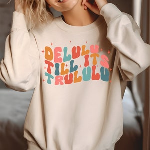 Puede incluir: Una sudadera beige con un estampado gr&aacute;fico colorido que dice "Delulu Till It's Trululu".