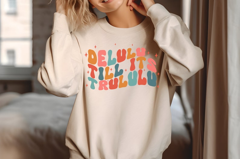 Puede incluir: Una sudadera beige con un estampado gr&aacute;fico colorido que dice "Delulu Till It's Trululu".