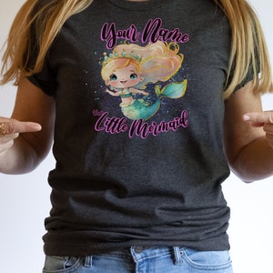 May include: A dark grey t-shirt with a cartoon mermaid illustration. The mermaid has blonde hair, a crown, and a turquoise tail. The text "Your Name the Little Mermaid" is printed on the shirt.