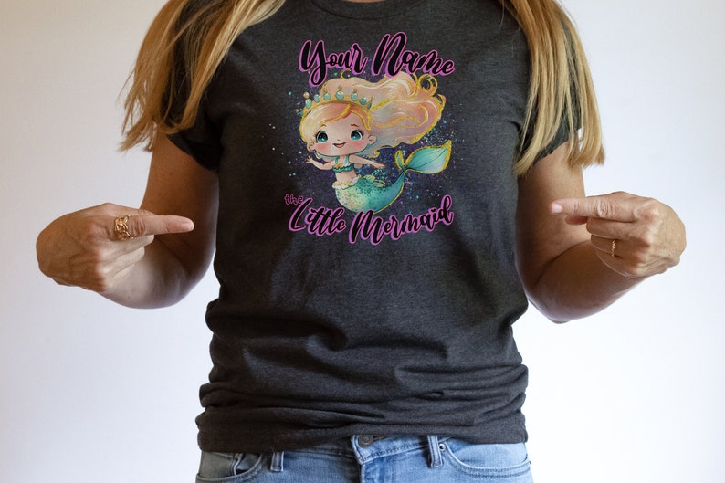 May include: A dark grey t-shirt with a cartoon mermaid illustration. The mermaid has blonde hair, a crown, and a turquoise tail. The text "Your Name the Little Mermaid" is printed on the shirt.