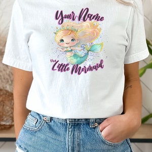 May include: White t-shirt with a cartoon mermaid wearing a crown and the text "Your Name the Little Mermaid" in purple.