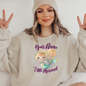 May include: A light beige sweatshirt with a graphic of a cartoon mermaid with a crown and the text "Your Name the Little Mermaid" in purple.