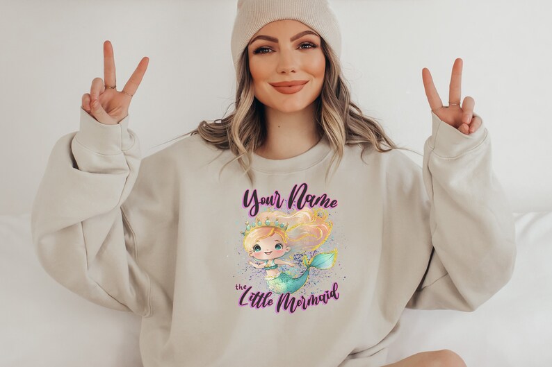 May include: A light beige sweatshirt with a graphic of a cartoon mermaid with a crown and the text "Your Name the Little Mermaid" in purple.