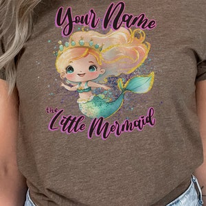 May include: A brown t-shirt with a cartoon mermaid wearing a crown and holding a seashell. The text "Your Name the Little Mermaid" is printed on the shirt.