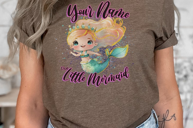 May include: A brown t-shirt with a cartoon mermaid wearing a crown and holding a seashell. The text "Your Name the Little Mermaid" is printed on the shirt.