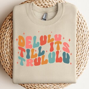 Puede incluir: Una sudadera beige con un dise&ntilde;o gr&aacute;fico colorido que dice "Delulu Till It's Trululu" con estrellas alrededor del texto.