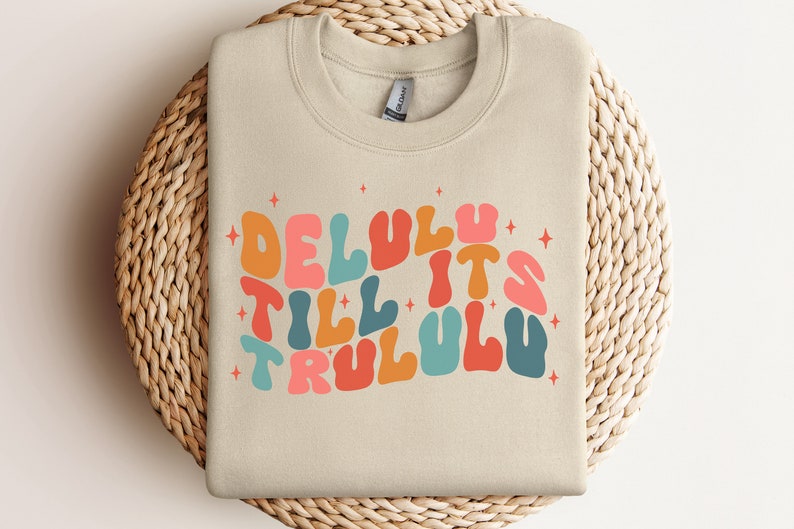 Puede incluir: Una sudadera beige con un dise&ntilde;o gr&aacute;fico colorido que dice "Delulu Till It's Trululu" con estrellas alrededor del texto.