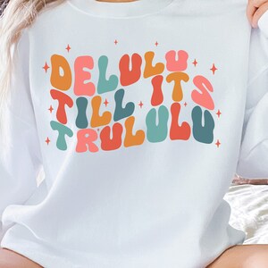 Puede incluir: Sudadera blanca con un gr&aacute;fico colorido que dice "Delulu Till It's TruluLu" en un estilo de fuente retro.