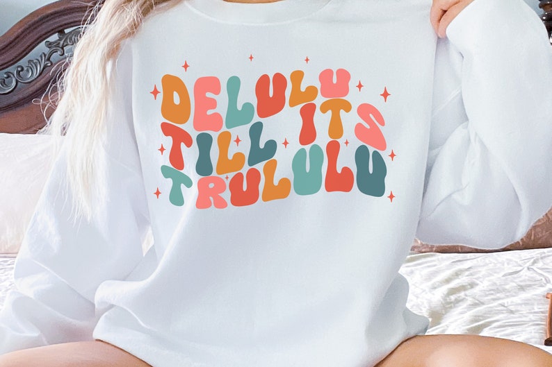 Puede incluir: Sudadera blanca con un gr&aacute;fico colorido que dice "Delulu Till It's TruluLu" en un estilo de fuente retro.
