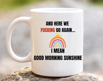 Pode incluir: Caneca de cerâmica branca com uma alça, com o texto "AND HERE WE FUCKING GO AGAIN... I MEAN GOOD MORNING SUNSHINE" em preto e vermelho, com um arco-íris e um sol.