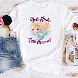 May include: White t-shirt with a mermaid illustration and the text "Your Name the Little Mermaid". The shirt is paired with denim shorts, a pink headband with black ears, white sneakers, and sunglasses.