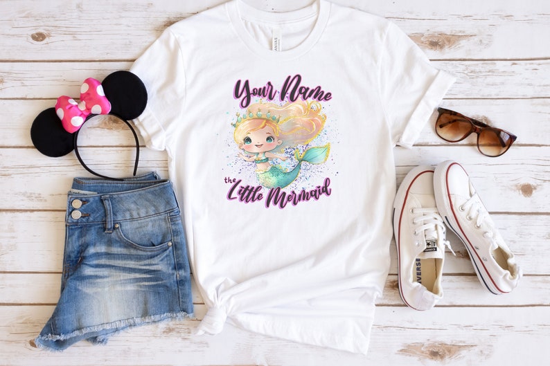May include: White t-shirt with a mermaid illustration and the text "Your Name the Little Mermaid". The shirt is paired with denim shorts, a pink headband with black ears, white sneakers, and sunglasses.