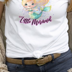 May include: White t-shirt with a watercolor illustration of a mermaid with blonde hair and a teal tail. The text "the little mermaid" is written in purple below the mermaid.