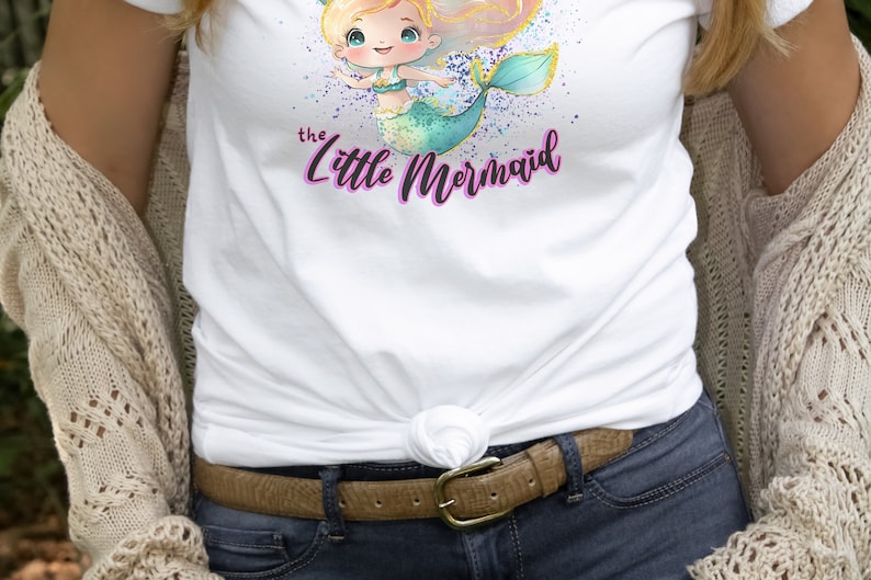 May include: White t-shirt with a watercolor illustration of a mermaid with blonde hair and a teal tail. The text "the little mermaid" is written in purple below the mermaid.