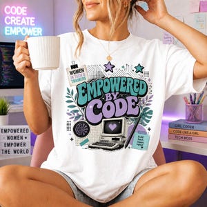 May include: A white t-shirt featuring a graphic print with the text "EMPOWERED BY CODE" in large teal and purple letters. The design includes a vintage computer with a heart on the screen, a brain icon, stars, leaves, and a note saying "build teach inspire repeat".