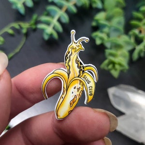 May include: A yellow and brown banana peel with a slug emerging from it. The slug has a yellow body with black spots. The text "SLUG CLUB" is on the banana peel. The image is held by a person.