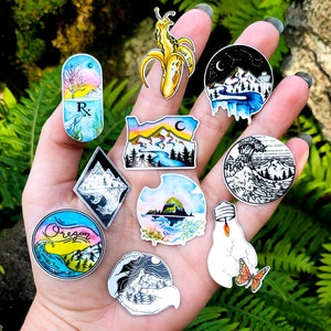 May include: A collection of colorful stickers with various designs, including a banana slug with "SLUG CLUB" text, mountain landscapes, a prescription pill with a tree, and a wave. The stickers are held in a hand against a backdrop of green foliage.