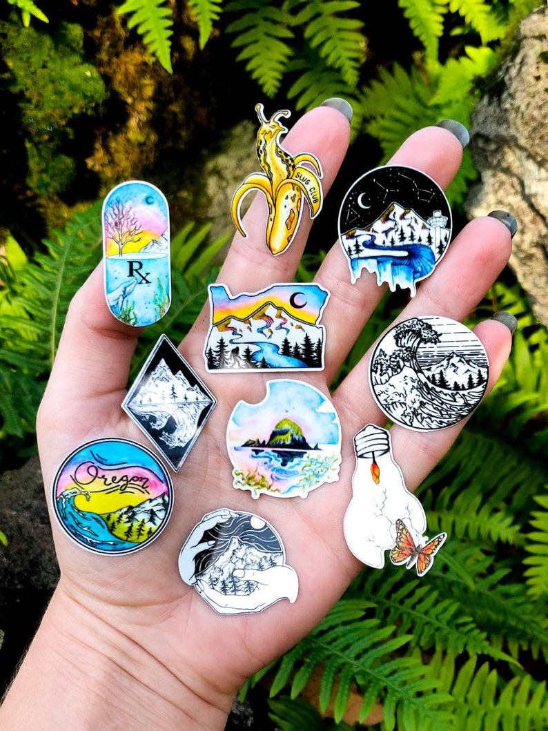 May include: A collection of colorful stickers with various designs, including a banana slug with "SLUG CLUB" text, mountain landscapes, a prescription pill with a tree, and a wave. The stickers are held in a hand against a backdrop of green foliage.
