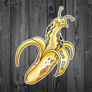 May include: A whimsical sticker featuring a yellow banana peel with a slug emerging from it. The slug has a patterned design and the text "SLUG CLUB" is on the banana peel. The sticker has a white border and is set against a dark wood background.