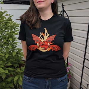 May include: A black short-sleeved t-shirt featuring a graphic print on the chest. The design displays a stylized red dragon with large wings positioned in front of a yellow flame-shaped background, creating a bold contrast against the black fabric.