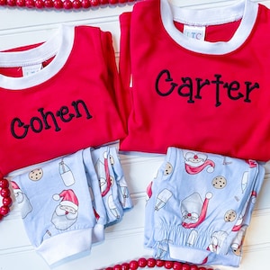 May include: Two sets of children's pajamas. The red long-sleeved shirts have white trim and black embroidered names. The light blue pajama pants feature a Santa Claus and cookie pattern. A string of red beads is in the background.