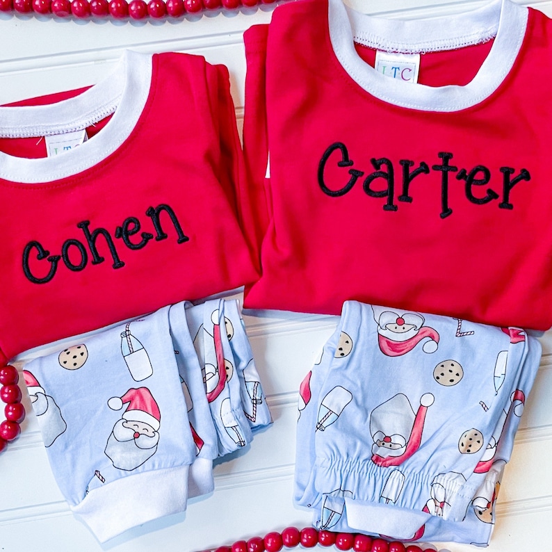 May include: Two sets of children's pajamas. The red long-sleeved shirts have white trim and black embroidered names. The light blue pajama pants feature a Santa Claus and cookie pattern. A string of red beads is in the background.