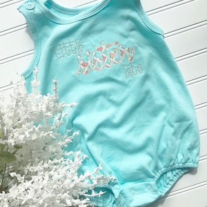 Könnte beinhalten: Ein hellblauer Baby-Strampler mit dem gestickten Text "Little Sister Tate" in rosa, weiß und grünem Karomuster.