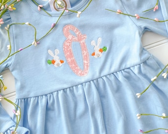 May include: A light blue long-sleeved dress with a pink embroidered letter "O" and two white bunnies holding carrots. The dress has a ruffled hem and is perfect for a spring celebration.