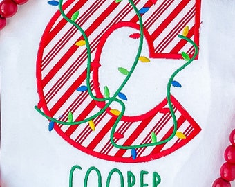 May include: White Christmas-themed shirt with a red and white striped letter "C" decorated with green and colorful lights. The name "COOPER" is embroidered in green below the letter.