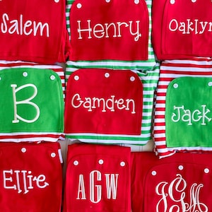 Puede incluir: Conjuntos de pijama personalizados rojos y verdes con nombres e iniciales bordados en blanco. Los pijamas tienen una parte superior roja con un bolsillo verde, o una parte superior verde con un bolsillo rojo. Los pantalones de pijama tienen rayas rojas y blancas.