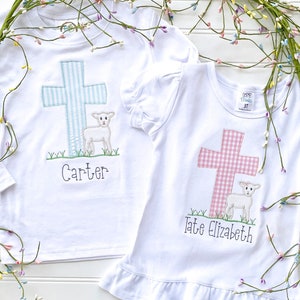 May include: Two white t-shirts with embroidered crosses and lambs. The cross on the left shirt is blue and white gingham, and the cross on the right shirt is pink and white gingham. The shirts are personalized with the names "Carter" and "Tate Elizabeth".