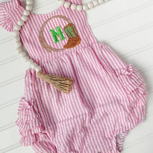 Puede incluir: Mono de beb&eacute; a rayas rosa y blancas con una letra M bordada en verde y marr&oacute;n y una bota en un c&iacute;rculo. El mono tiene volantes en las mangas y alrededor de las piernas. Un collar de cuentas blancas con un borla marr&oacute;n est&aacute; en el fondo.
