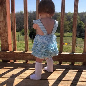 Puede incluir: Una ni&ntilde;a peque&ntilde;a con un vestido a rayas azules y blancas con volantes y calcetines blancos hasta la rodilla se para en una plataforma de madera mirando hacia un campo de hierba.