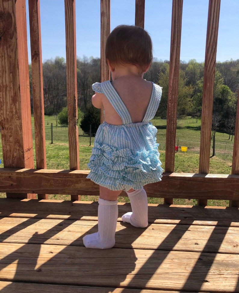 Puede incluir: Una ni&ntilde;a peque&ntilde;a con un vestido a rayas azules y blancas con volantes y calcetines blancos hasta la rodilla se para en una plataforma de madera mirando hacia un campo de hierba.