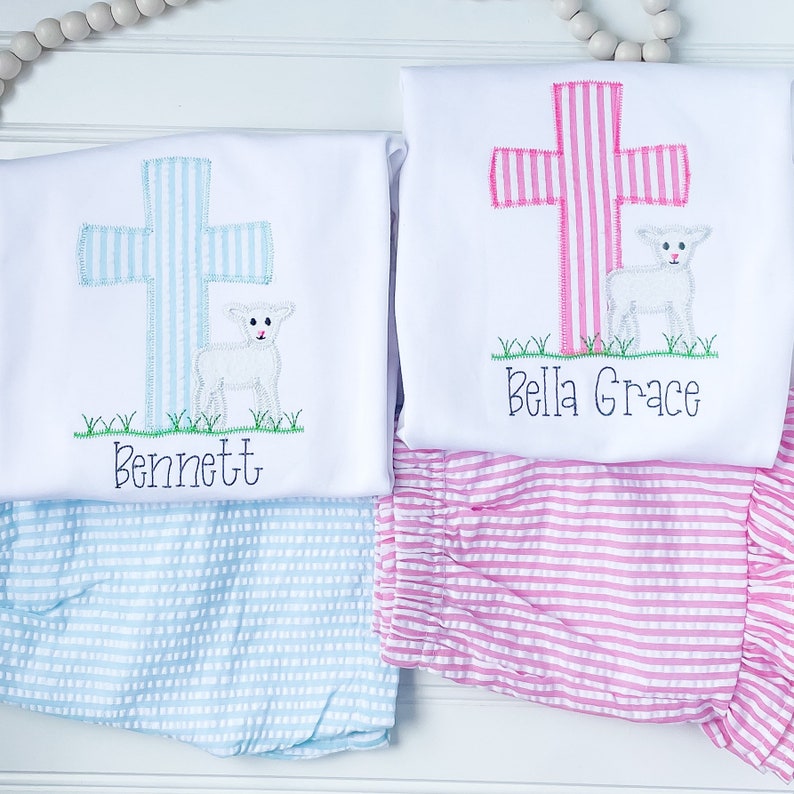 May include: Two white onesies with embroidered crosses and lambs. The onesies are personalized with the names "Bennett" and "Bella Grace".