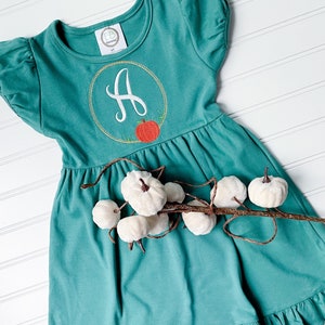 May include: Teal-colored dress with short sleeves and a ruffled hem. The dress features an embroidered gold-outlined circle with the letter "A" and a small orange pumpkin. A decorative branch with white pumpkins is in the foreground.