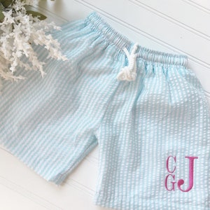 Puede incluir: Bañador de rayas azul claro y blanco con cordón blanco y iniciales bordadas en rosa "C G J".