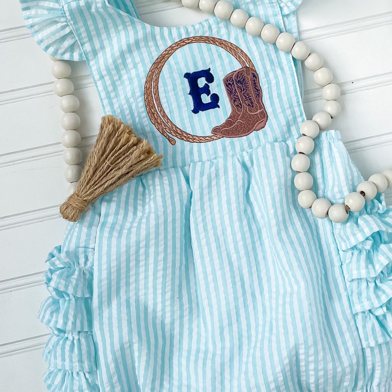 Puede incluir: Un pelele de beb&eacute; a rayas azul claro y blanco con un dobladillo con volantes. El pelele tiene un dise&ntilde;o bordado de una bota de vaquero y un lazo con la letra "E" en el centro. Un collar de cuentas blancas est&aacute; drapeado sobre el pelele.