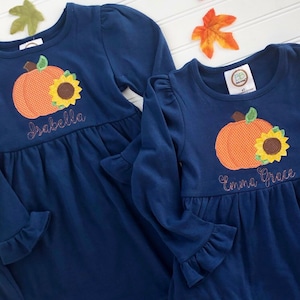 May include: Two navy blue long-sleeved dresses with pumpkin and sunflower embroidery. The dress on the left says "Isabella" and the dress on the right says "Emma Grace".