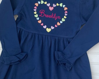 Puede incluir: Un vestido azul marino de manga larga con un diseño en forma de corazón hecho de corazones coloridos. El nombre "Brooklyn" está bordado en rosa dentro del corazón.