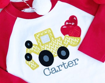 Puede incluir: Una camiseta de estilo béisbol blanca y roja con un vehículo de construcción amarillo que lleva corazones rojos. La camiseta está personalizada con el nombre "Carter" en gris.
