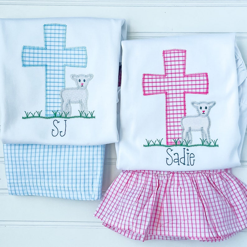 K&ouml;nnte beinhalten: Zwei wei&szlig;e Hemden mit gestickten Kreuzen und L&auml;mmern. Das Hemd links hat ein blaues Kreuz und den Text "SJ". Das Hemd rechts hat ein pinkfarbenes Kreuz und den Text "Sadie".